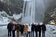 A group of 10 students stand on a glacier in front of a waterfall.
