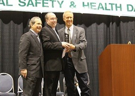 Illinois Department of Labor Director Hugo Chaviano (left) and Gov. Bruce Rauner (right) present Illinois State alum John Lee with the Governor’s Award for Contributions in Health and Safety.