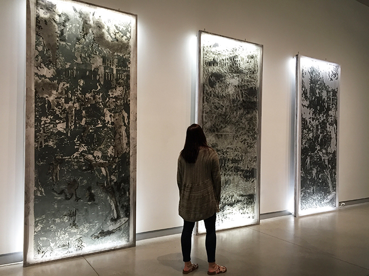 See a woman from behind as she walks past three 12-foot tall multi-layered prints that are lilluminated by florescent lights.
