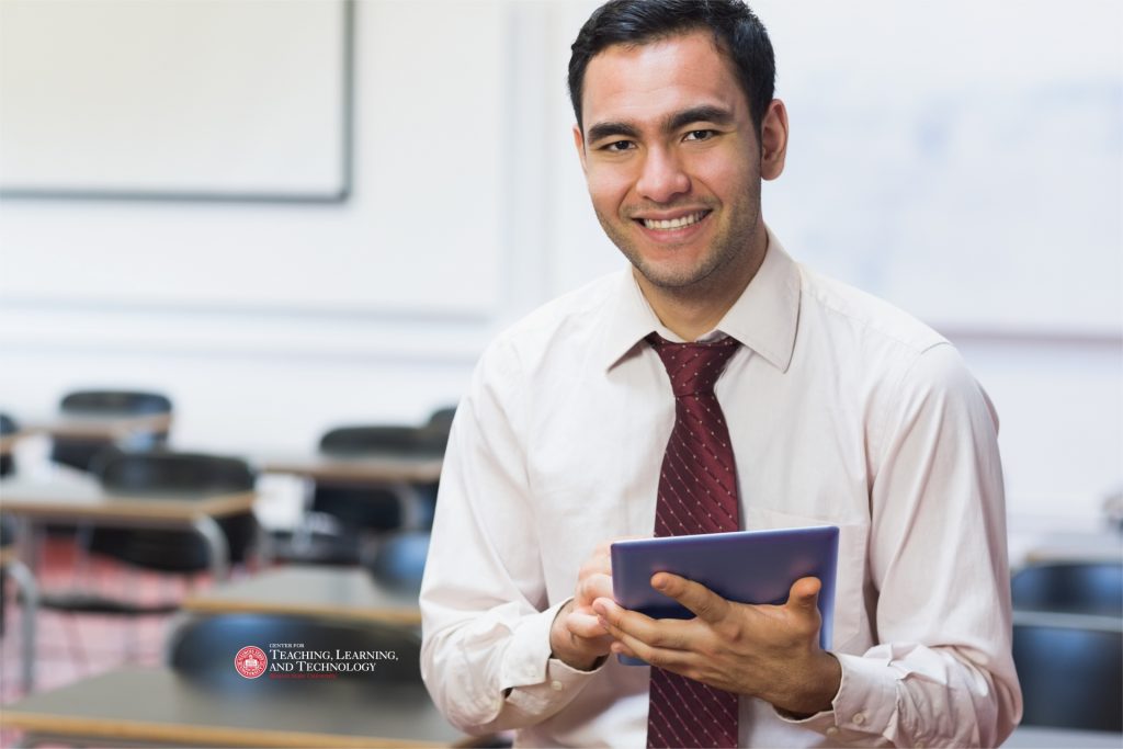 Instructor in empty class, smiling into the camera, holding a tablet device.