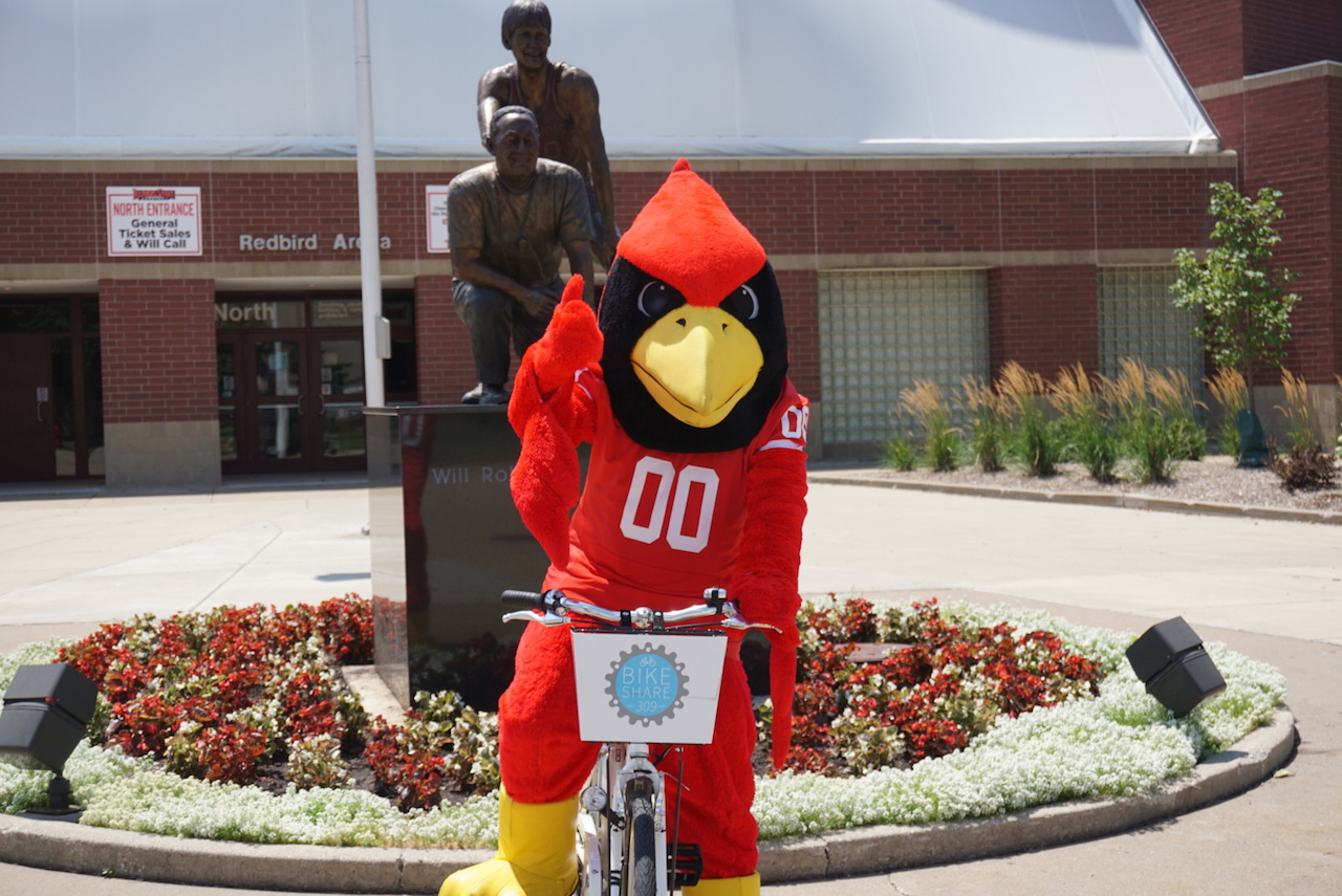 Reggie Redbird rides a bike from the Town of Normal's rental program, Bike Share 309.
