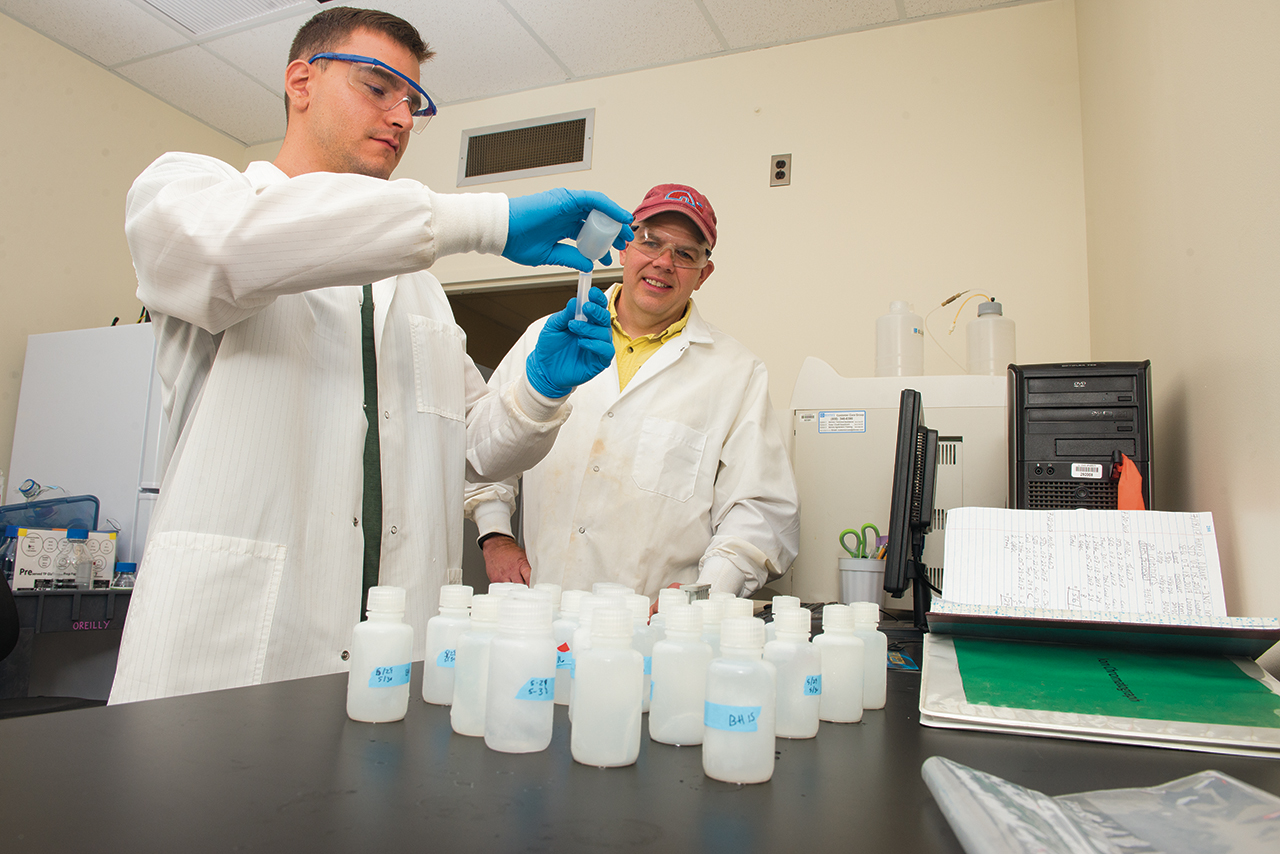 two men in lab coats, one pouring a substance into a beaker