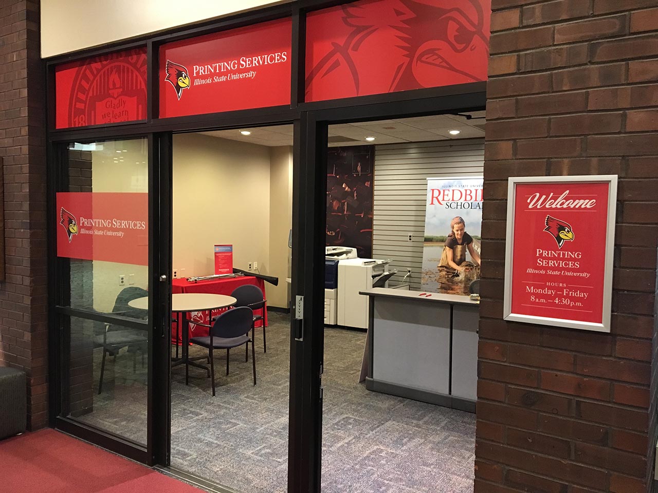 The open, glass doors of the Printing Services Office, with a red banner and a desk.