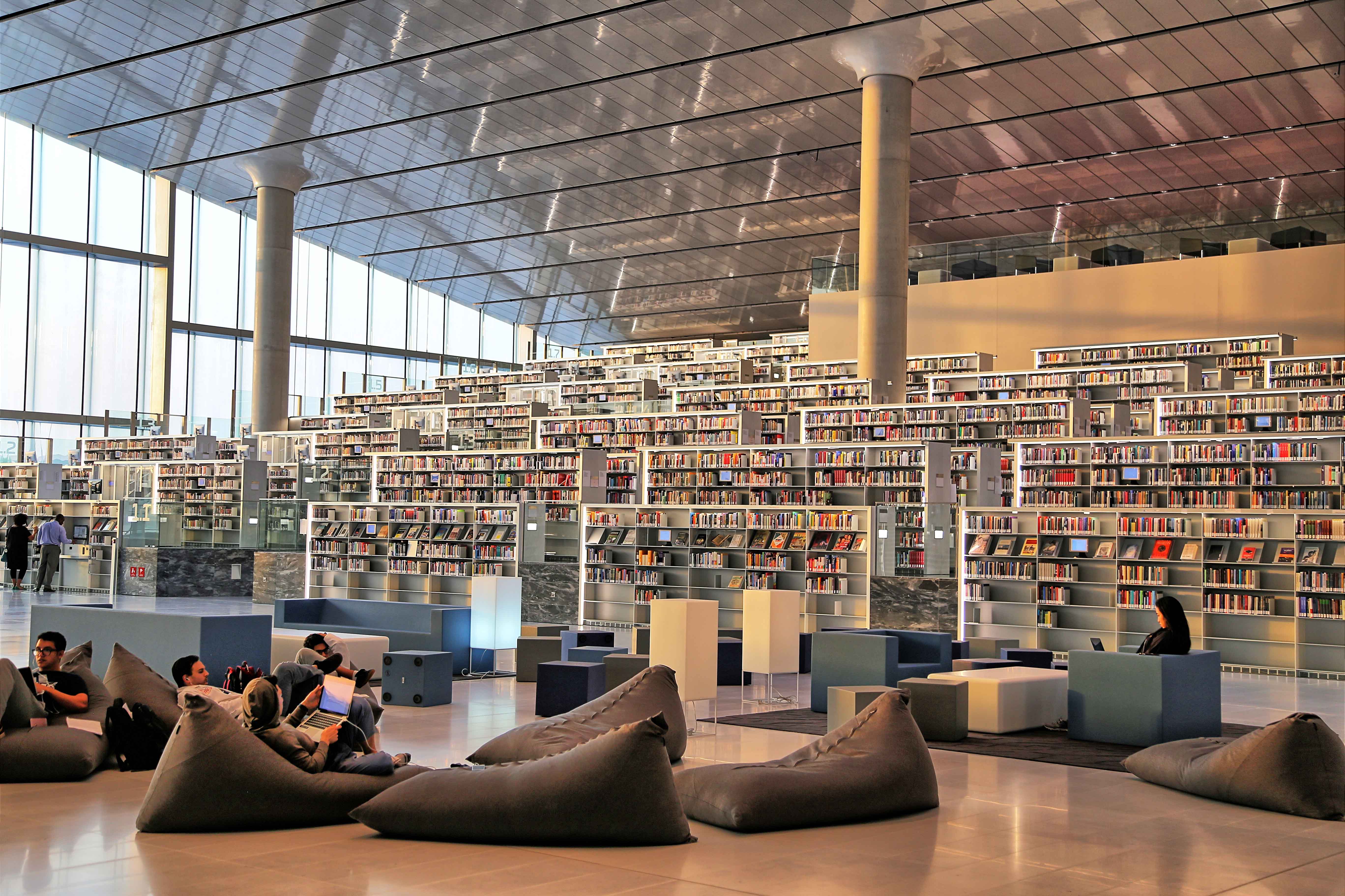 Tiered rows of stacks of books set against a vaulted ceiling of concrete and glass. People in large bean bangs in the foreground.