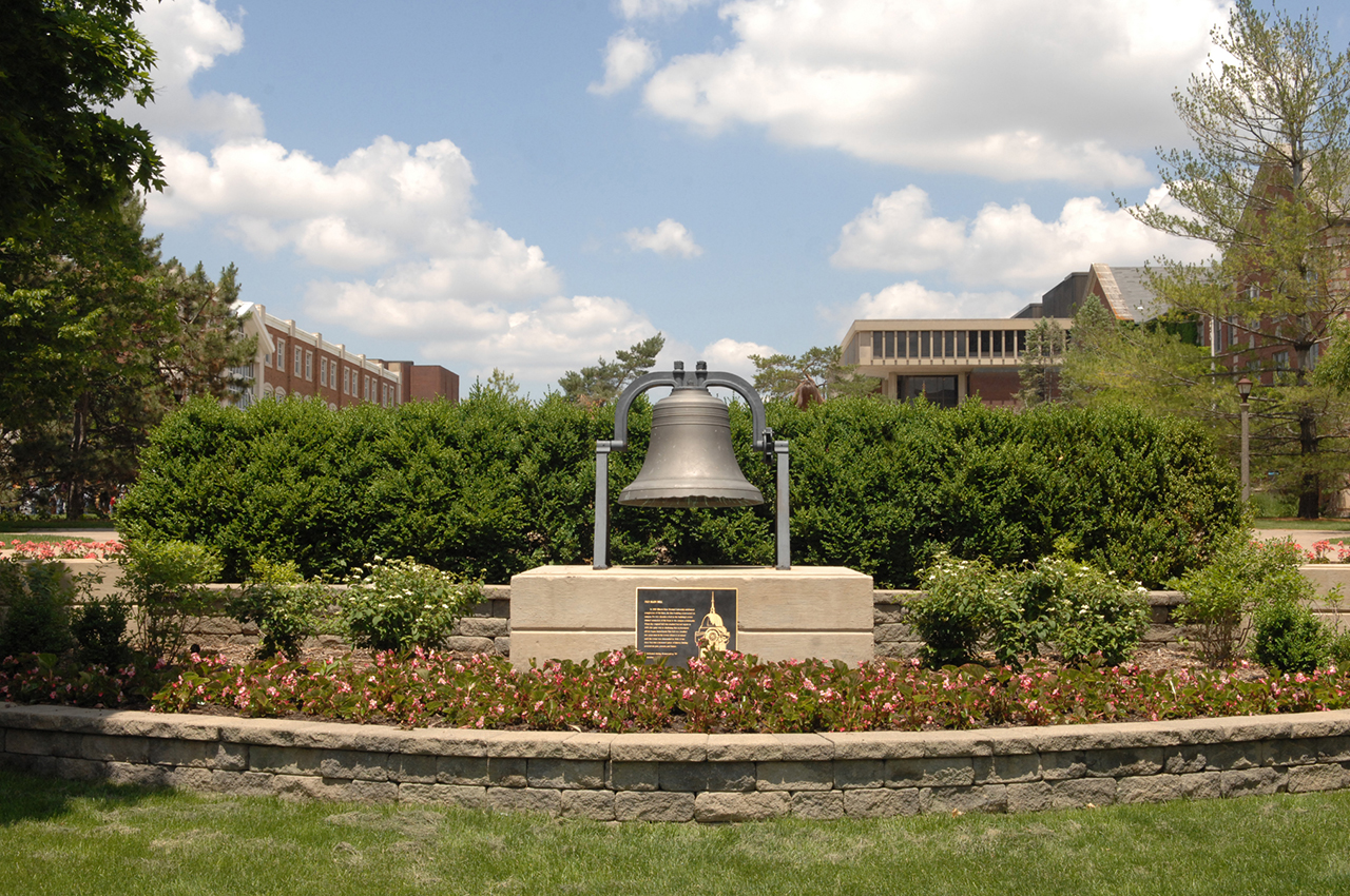 Large bell surrounded by flowers and set in stone landscaping, with academic buildings in the background
