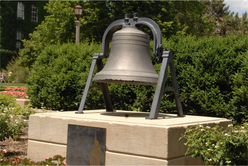 Large bell surrounded by flowers and set in stone landscaping, with trees and academic buildings in the background