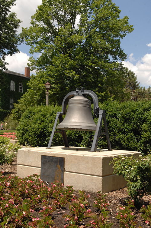 Large bell surrounded by flowers and set in stone landscaping, with trees in the background