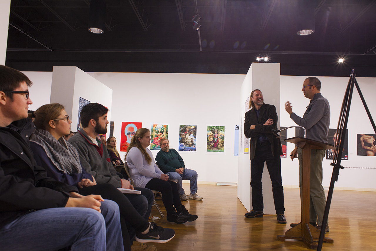 Two men stand a podium and address an audience seated in an art gallery.