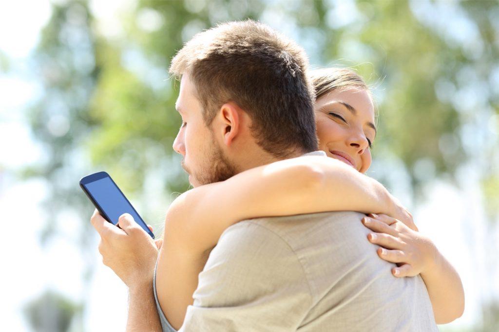 Man and woman embracing, but man looking at his cell phone