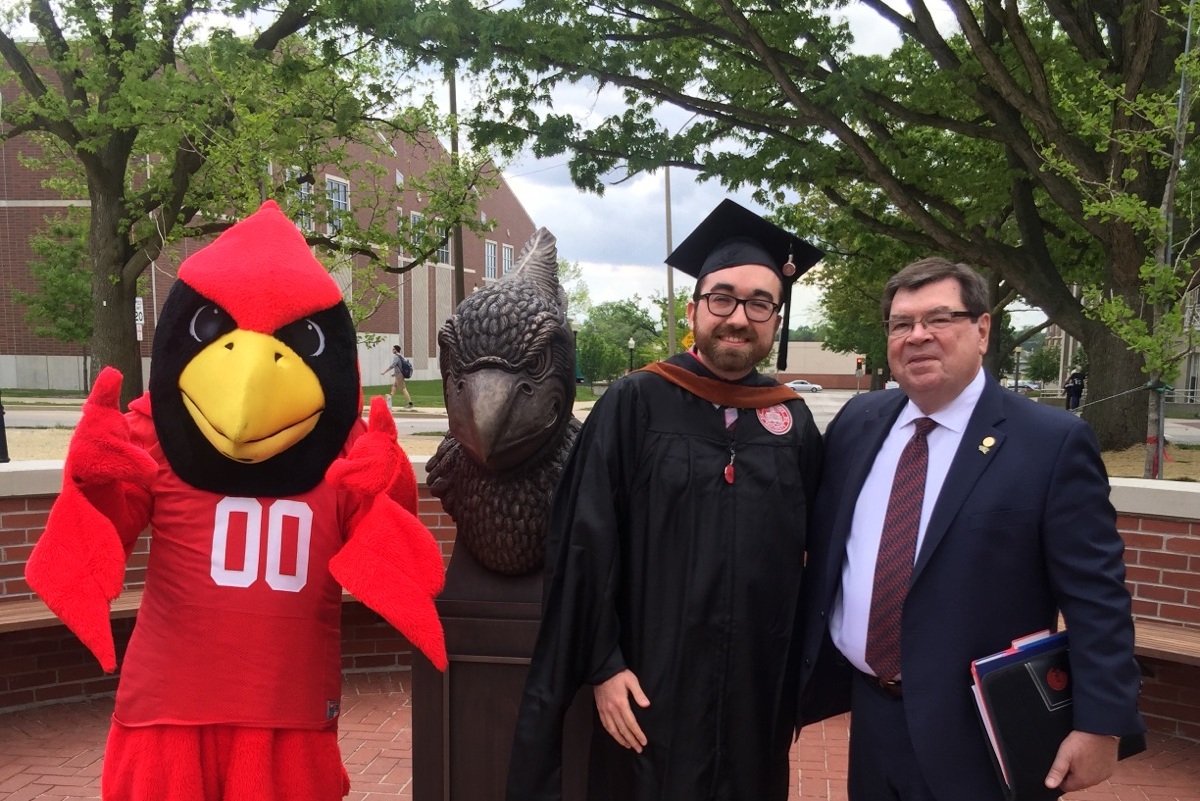Reggie Redbird, student in cap and gown, and President Dietz posing in Redbird Plaza