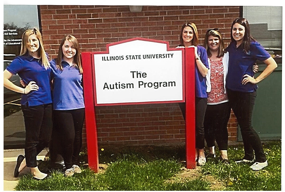 4 graduate students and 1 instructor stand next to a sign for Illinois State University The Autism Program