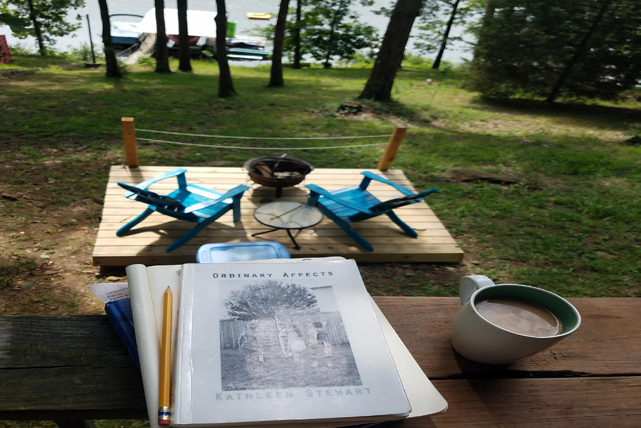 Image taken during the residency of the lake, a boat dock, a patio with a fire pit, books about art, and a coffee mug.