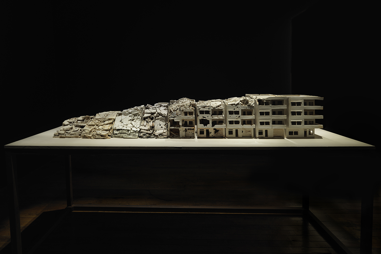 Photograph of art installation by Alison Ruttan depicting a series of buildings in various states of destruction from complete destruction to fully intact.
