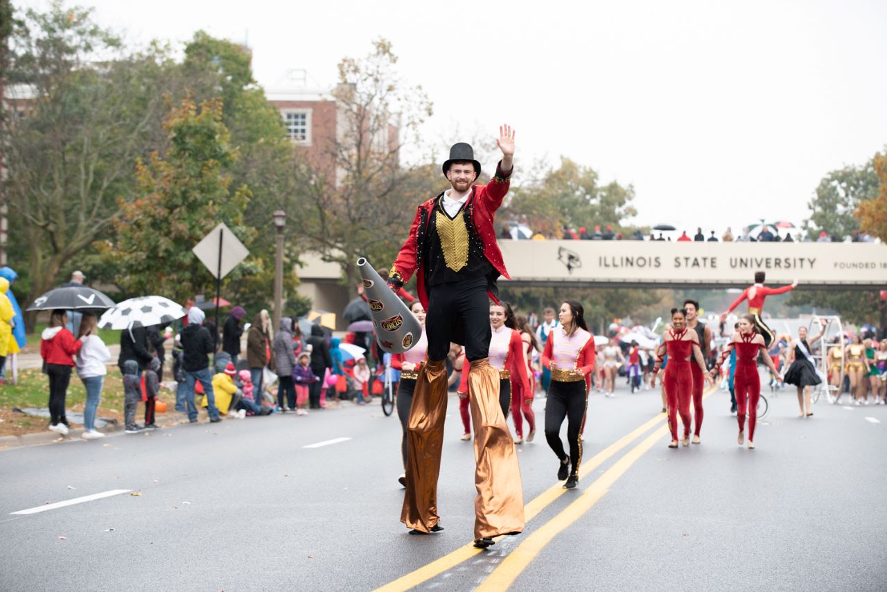 Ringmaster on stilts waving in 2019 ISU Homecoming Parade with performers in the background.