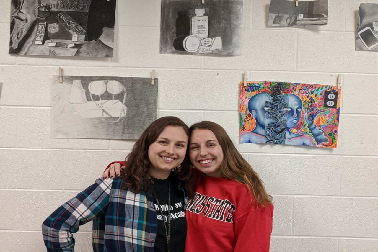 Two teachers pose in front of student art projects