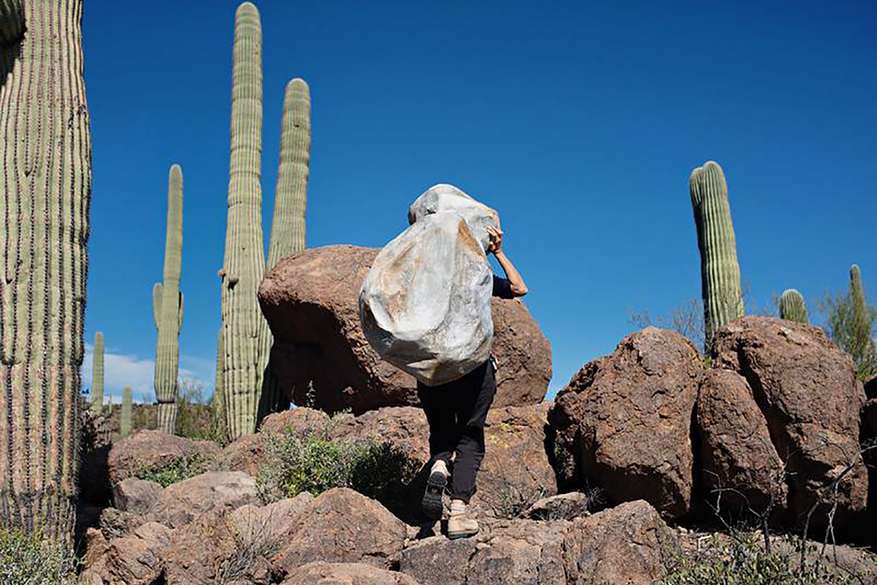 Nazafarin Lotfi, Transit, 2020. Digital photograph. Photo shows a person walking among cacti and boulders, carrying a sculptured boulder on their back.