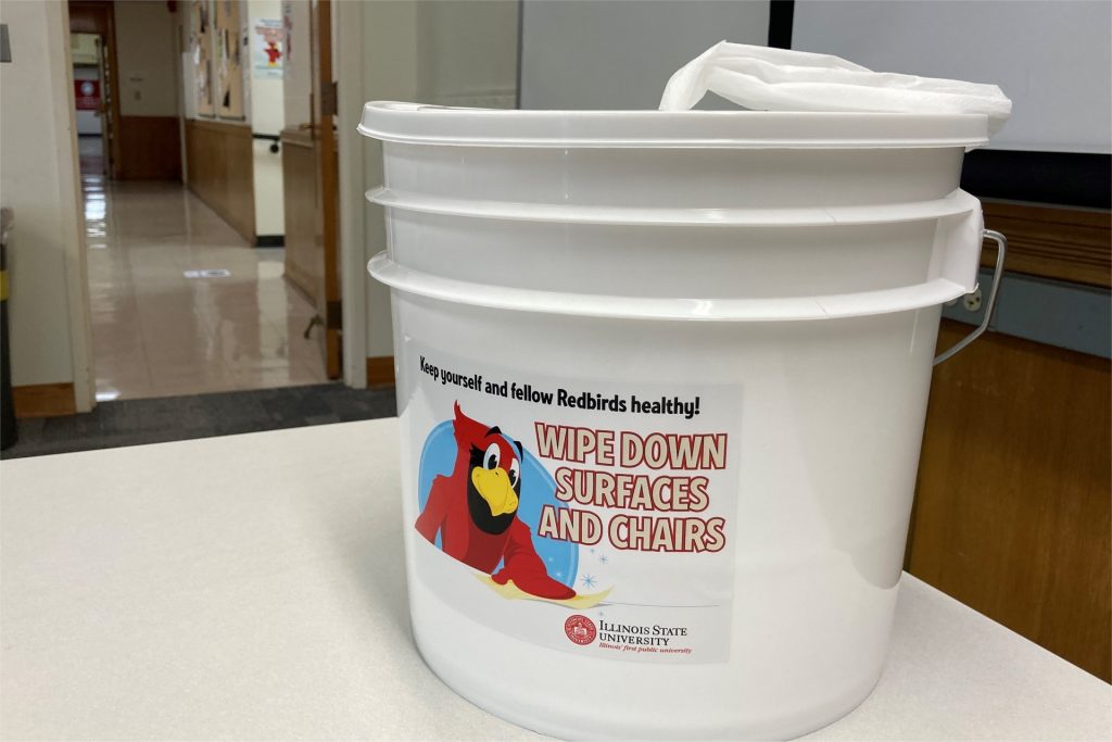 A tub of disinfectant wipes in a classroom, featuring Reggie Redbird and the text, "Keep yourself and other Redbirds healthy! Wipe down surfaces and chairs."