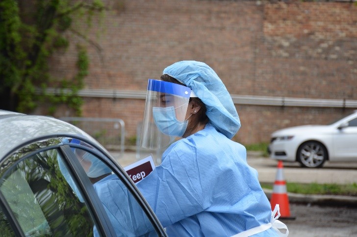 Woman in nurse suit, face shield, and face mask reaches into car at testing site