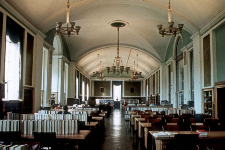 Photograph of Milner LIbrary's Reading Room in 1971 when the library was in what is now Williams Hall. ROom has very high ceiling, ornate fixtures, open tables, and is surrounded by books.