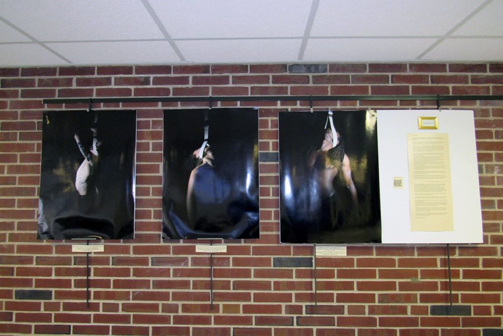 A picture of 3 large photos of Black circus performers hanging from circus straps from their heads and a 4th poster of a written explanation of this "Strange Fruit"