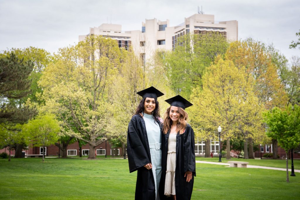 Two women wear commencement caps and gowns pose on the green Quad with Watterson in the distance behind them