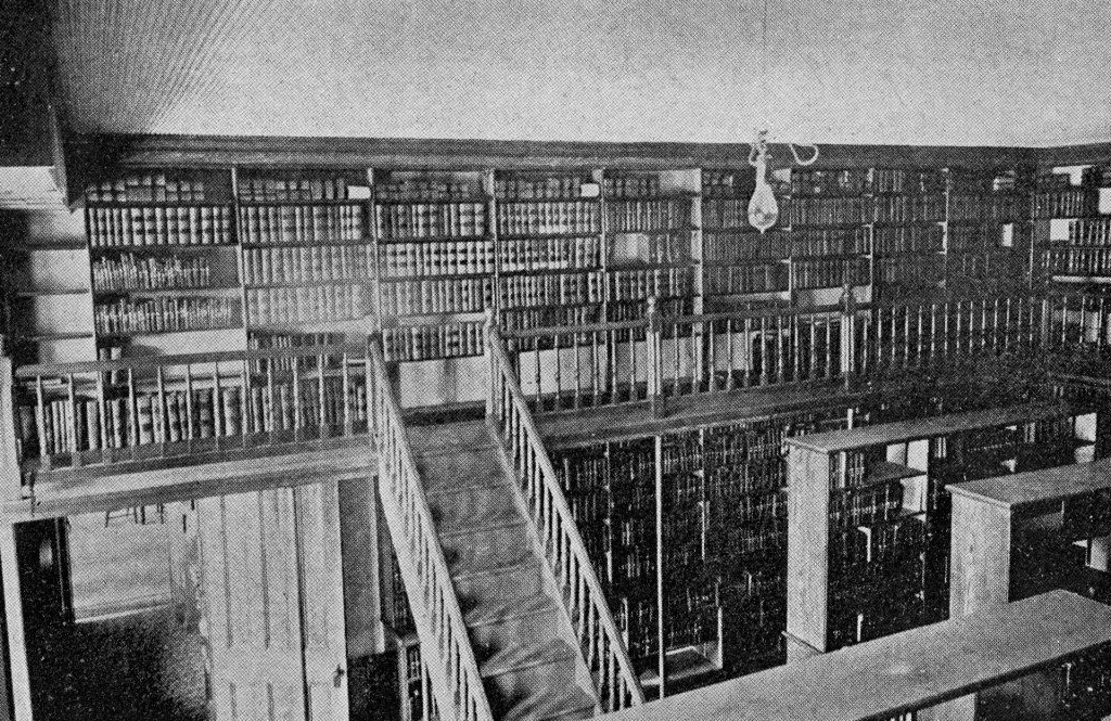Black and white photograph of the University's library when it was Old Main consisting of a room with books on shelves and then a second floor balcony area with a wall of bookshelves. 