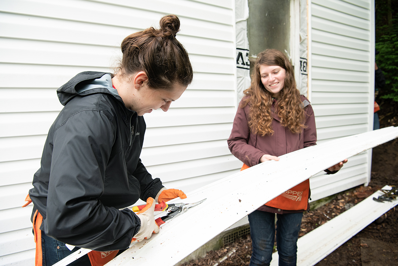 Katie Dowling (right) holds a piece of siding level as William Restis (left) uses a pair of tin snips to trim it down to fit around the under hang of the house.