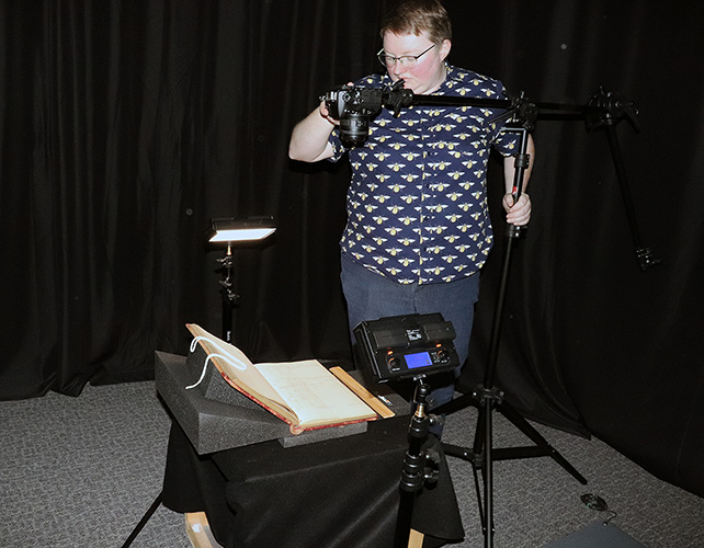 Person uses camera on a tripod and lighting apparati to photograph an old book for digitization. 