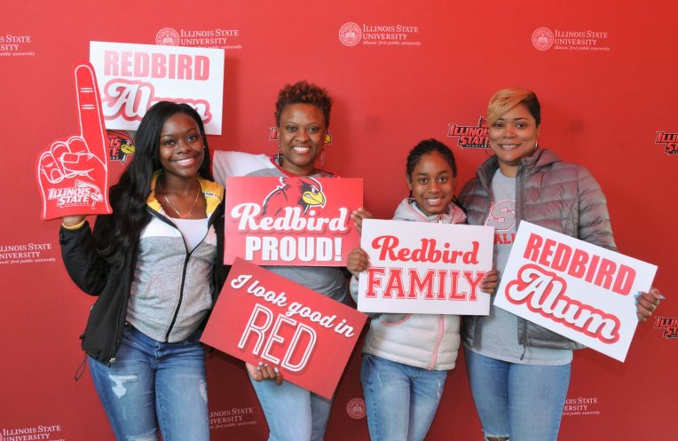 A Redbird family posing with spirit signs in front of a red Illinois State background