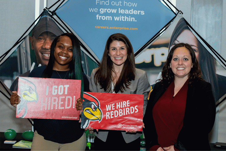 student holding the "I Got Hired" sign as her employer holds the "We Hire Redbirds" sign