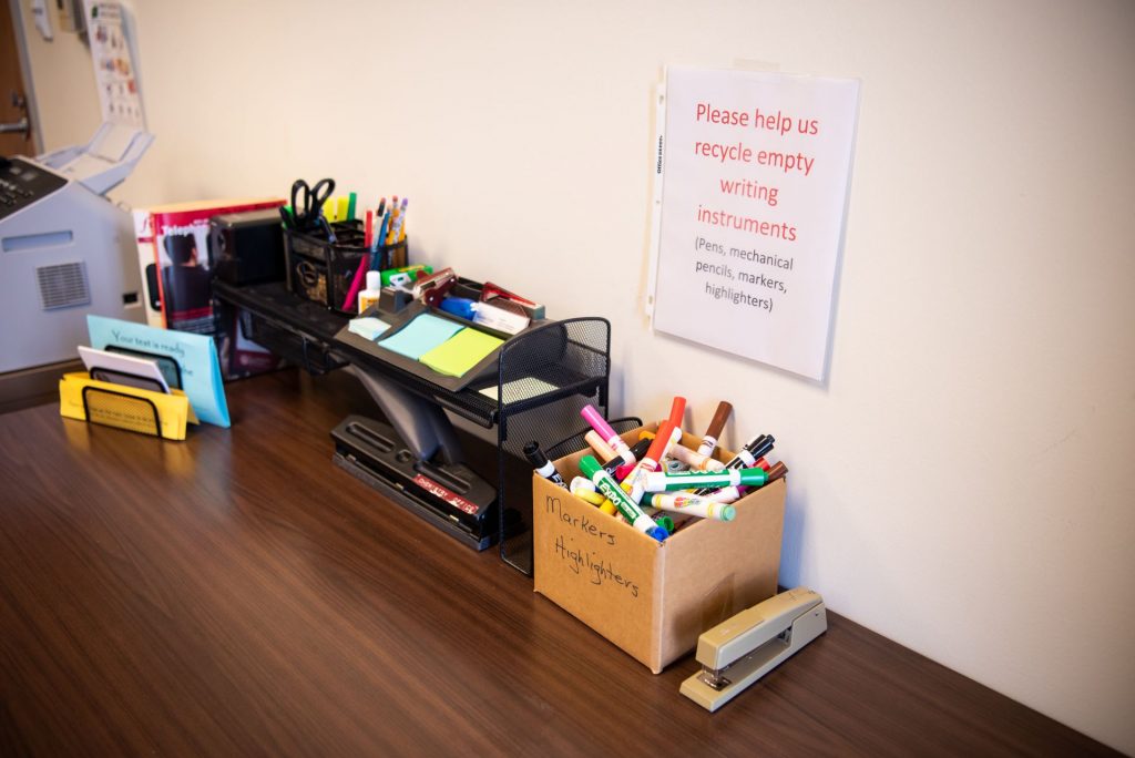 A box labeled "Markers + Highlighters" is full of writing utensls, and sits under a sign that reads "Please help us recycle empty writing instruments."