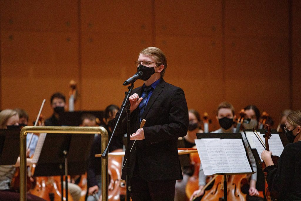 Assistant Symphony Orchestra conductor Matthew Clarke stands at the mic in front of the orchestra.