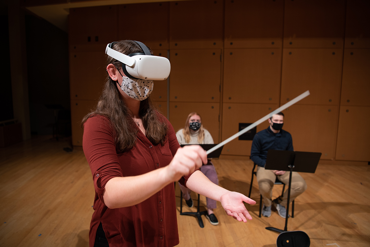 One student stands in the foreground, wearing a VR headset and holding up a conducting baton. Two students sit in the background behind her.