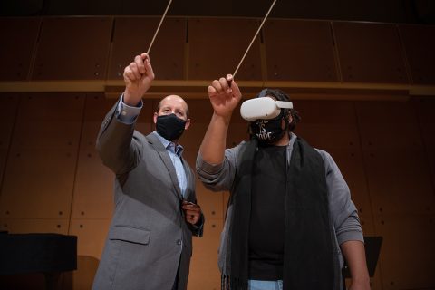Dr. Tony Marinello stands next to a student. The student is wearing a VR headset and both of them are holding up a conducting baton.