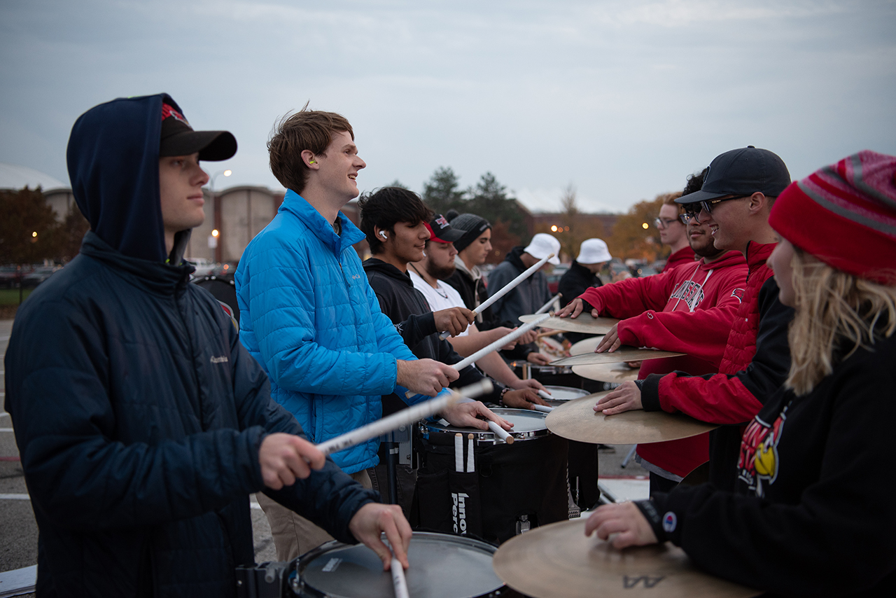 Rudy Morr smiles as he drums with other band members. He stands beside other students playing the snare and across from a line of students playing the cymbals.