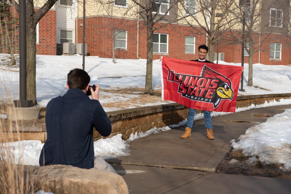 Victor Ventura stands in the Cardinal Court courtyard holding an Illinois State wall flag. Simon Schuler kneels in front of him with a camera to take his picture.