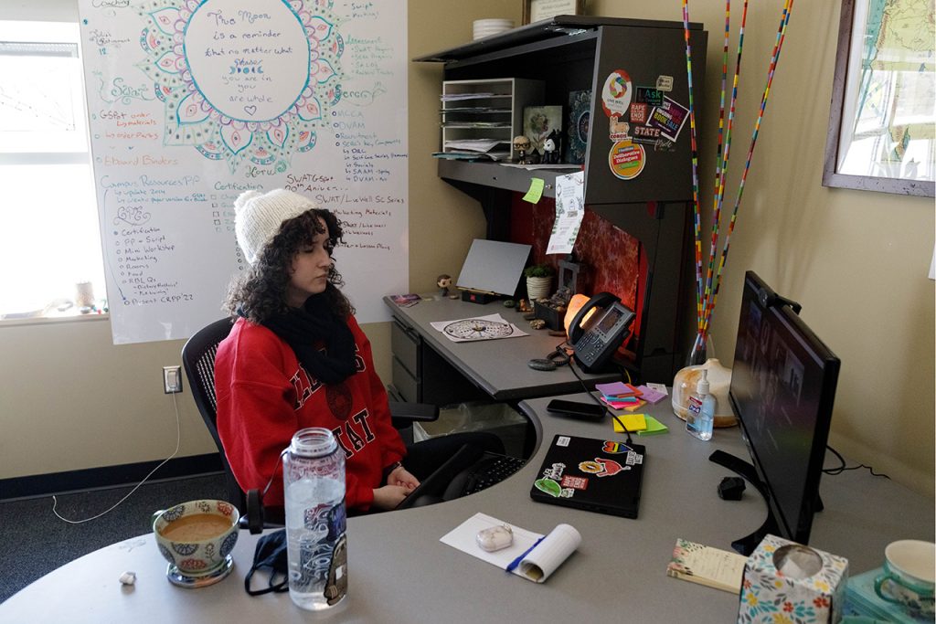 Misia Grzybowski sits in her office with her eyes closed meditating. In front of her is a desktop computer, on which is the Zoom Koru Mindfulness class.