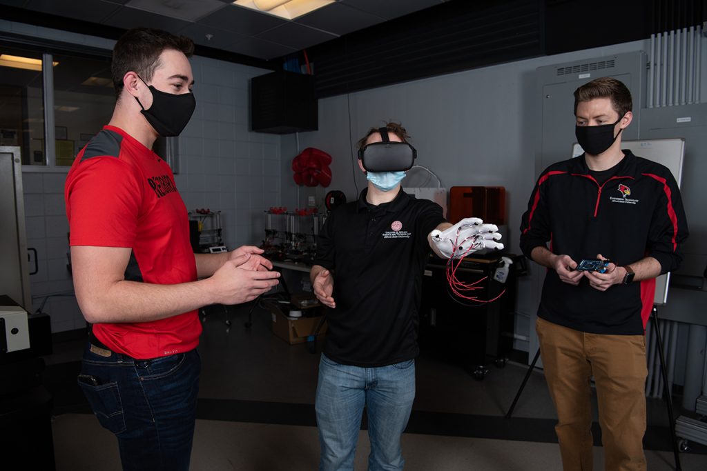 Jake Weihe stands on the far left. Alex Diffor stands in the center, wearing a VR headset and a haptic glove on one hand, which is extended out in front of him. Jordan Osborne stands on the right, holding a small circuit board.