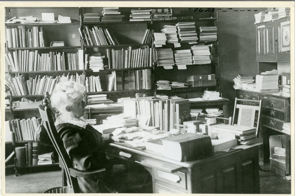 A black and white vintage photo of a woman sitting in a chair surrounded by books.