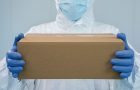 Person in surgical gloves, outfit, mask holding a cardboard box