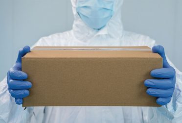 Person in surgical gloves, outfit, mask holding a cardboard box