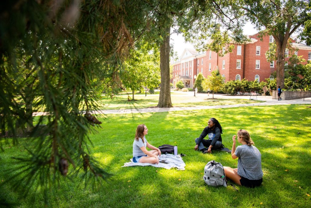 Three students sit in a circle on the green grass of the Quad. Fell Hall is in the background with pine tree branches creeping into the photo frame in the foreground.