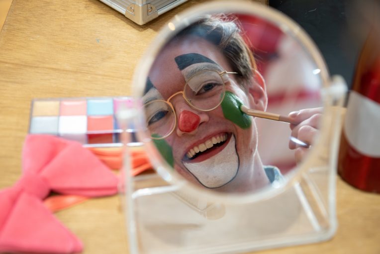 Photo of Jon Fulton's reflection in a mirror. Fulton is wearing clown makeup and using a makeup brush to finish a green circle on his cheek.