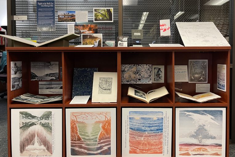 Exhibit with books and prints laid out along the top of a brown display case. Eight square openings below hold books, photographs, and prints.