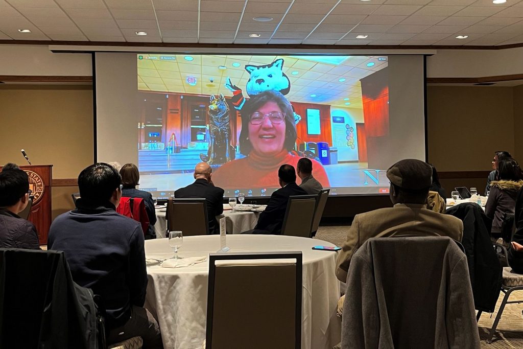 woman speaking on a video screen to a room of people
