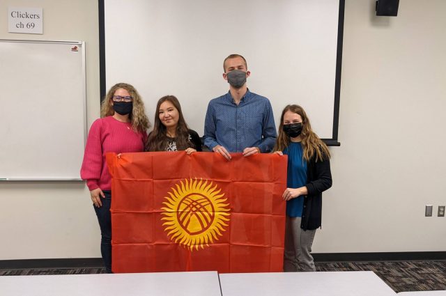 A faculty member and three students hold a flag at the front of a classroom.