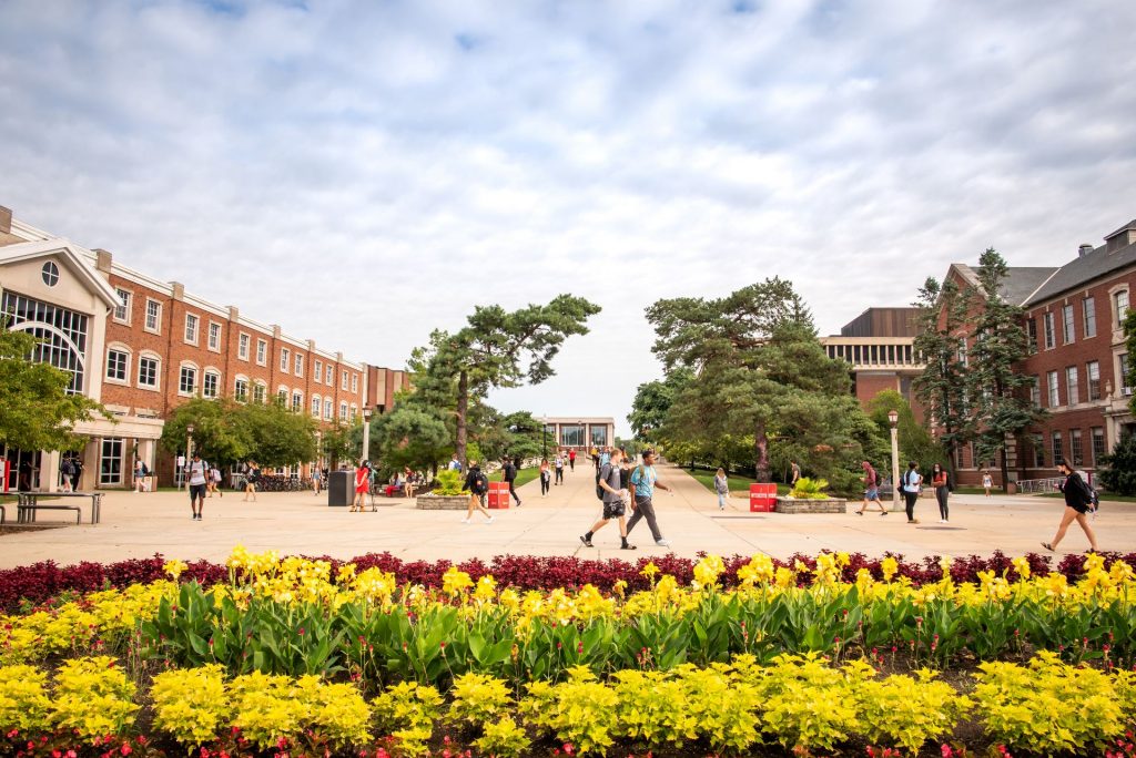 Image of students walking across the Quad with flowers in the foreground, Schroeder Hall on the left, Felmley Hall on the right, and trees archiing over the Quad walkway bridge in the middle of the background.