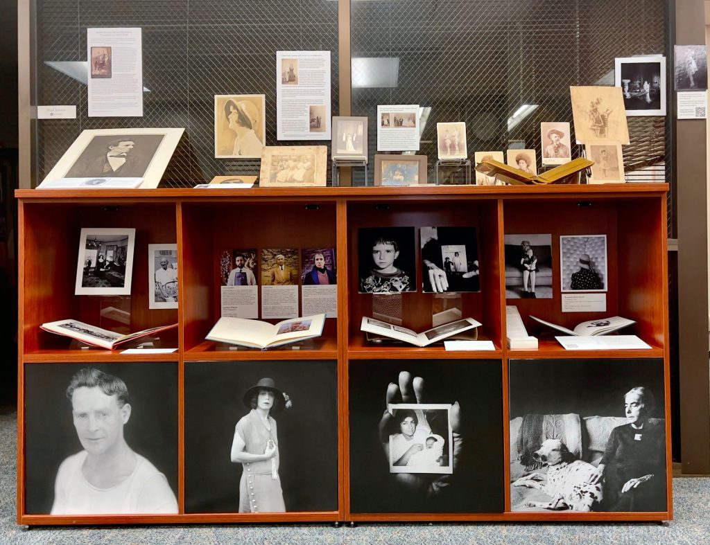 : Exhibit case with eight square sections filled with photographs and books. All images are portraits