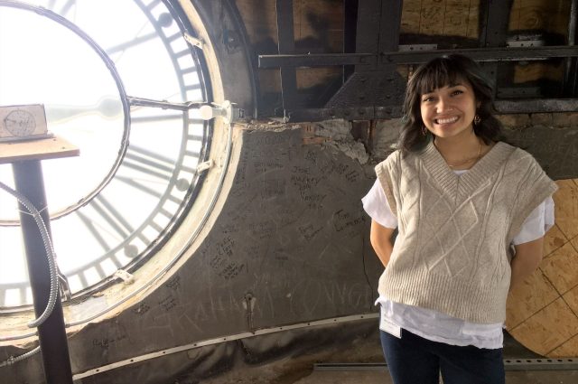 Woman standing next to a the face of a large clock inside the top of an old courthouse