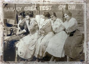 Five women seated in a line in front of a circus wagon that reads Bailey Greatest Show. The women are wearing ankle-length skirts and dresses and are posed with their hands resting on the shoulders of the woman next to them